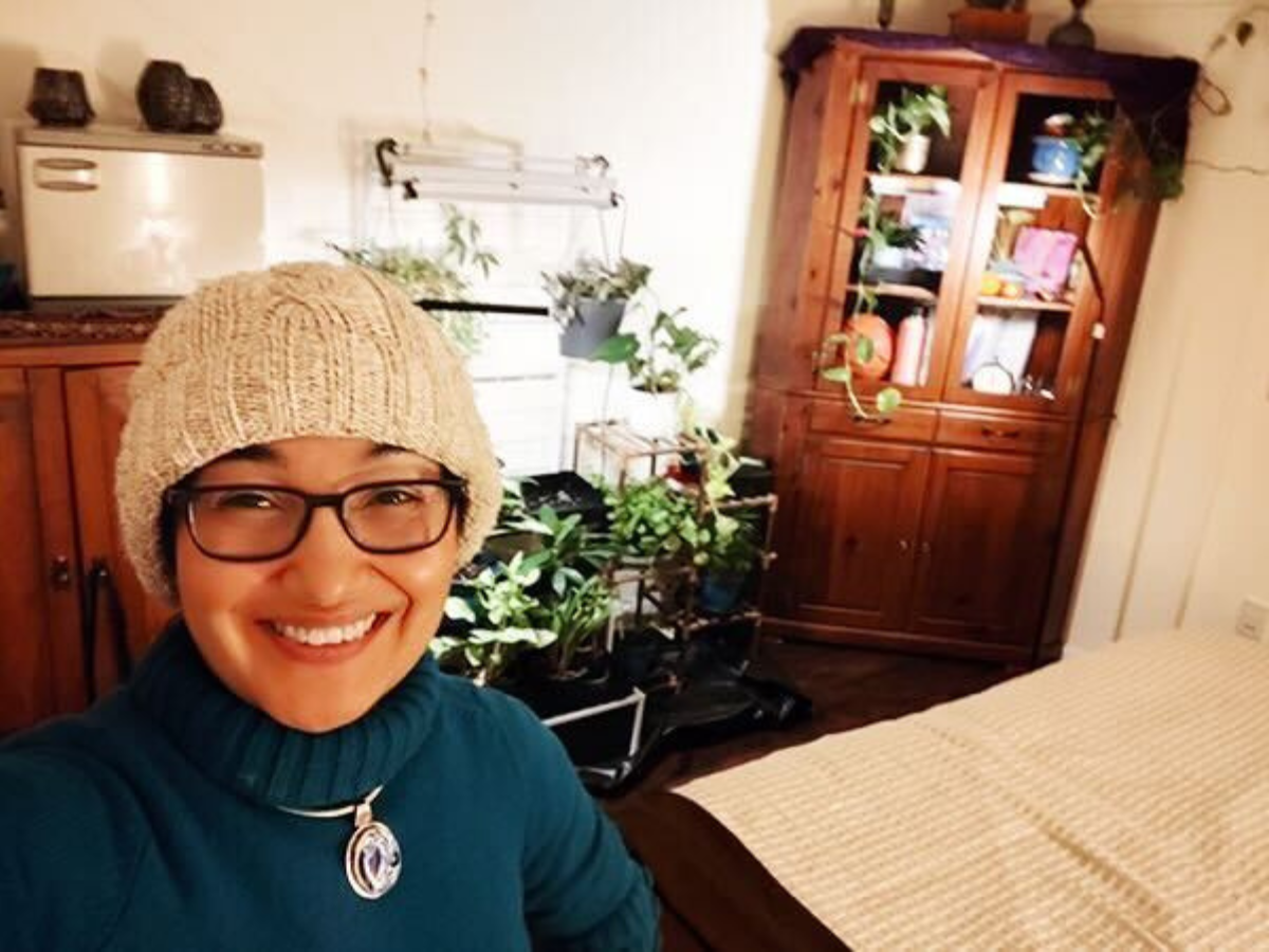 A person wearing a knit hat and glasses smiles at the camera in a room with a bed, wooden furniture, and several potted plants.