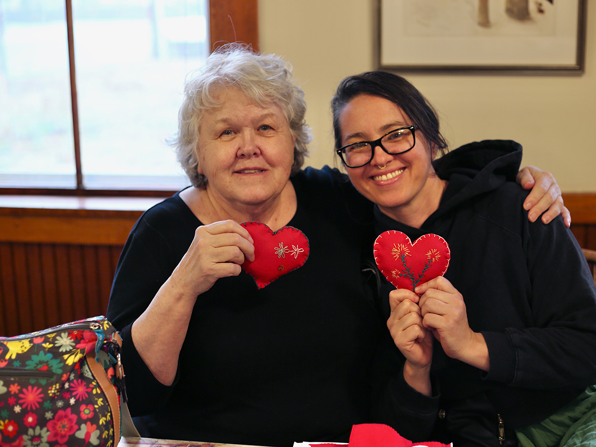 Two people smiling, sitting close together, and holding red heart-shaped crafts with embroidered details. A colorful bag is on the table in front of them.