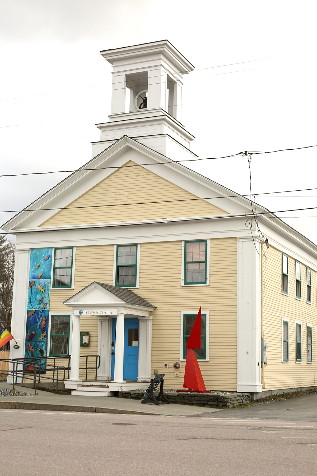 A two-story building with a bell tower, yellow siding, and white trim houses the River Arts gallery. Two colorful murals and a red sculpture decorate outside. Power lines overhead.