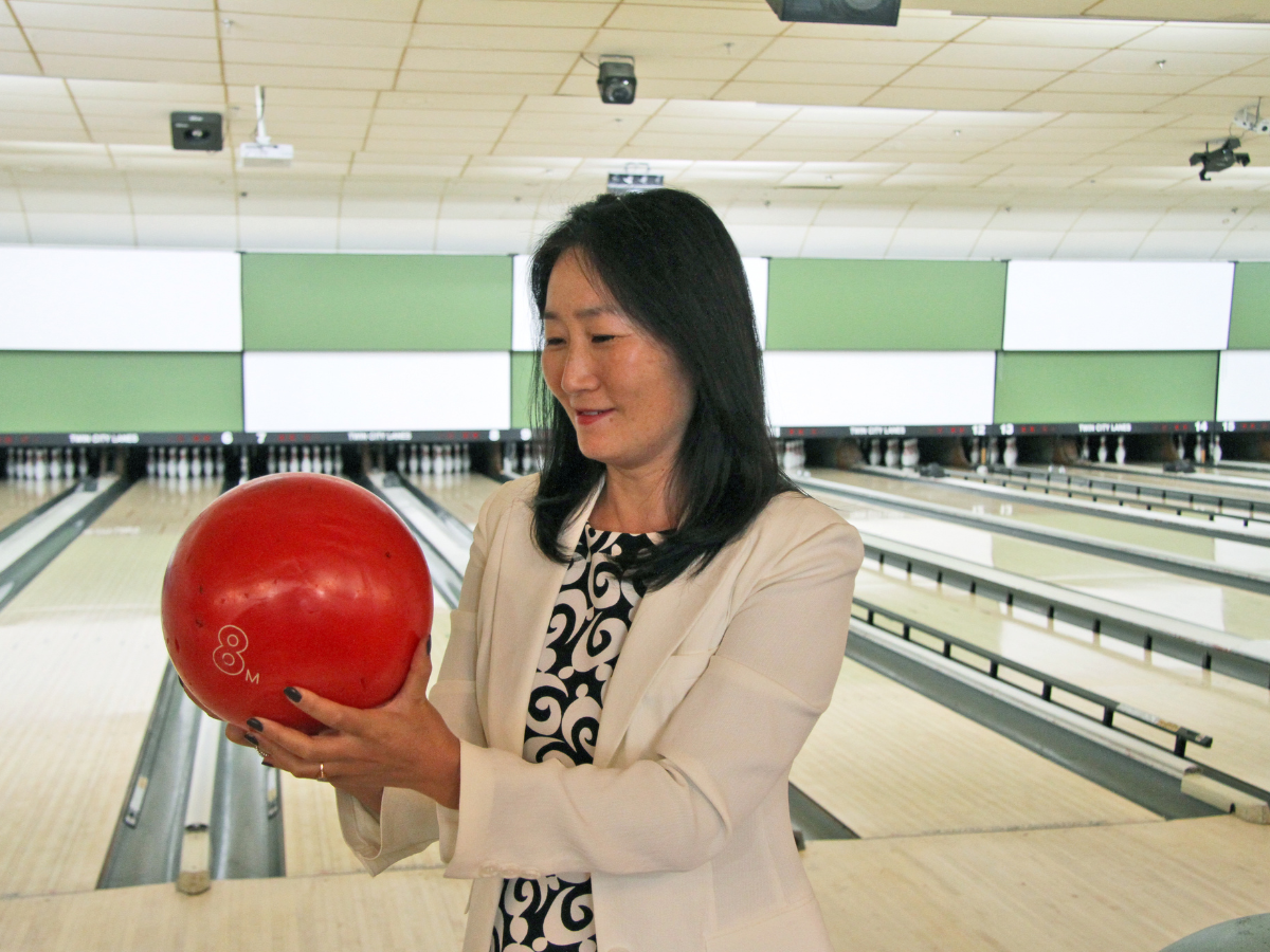 A woman in a white jacket and patterned dress holds a red bowling ball at a bowling alley.