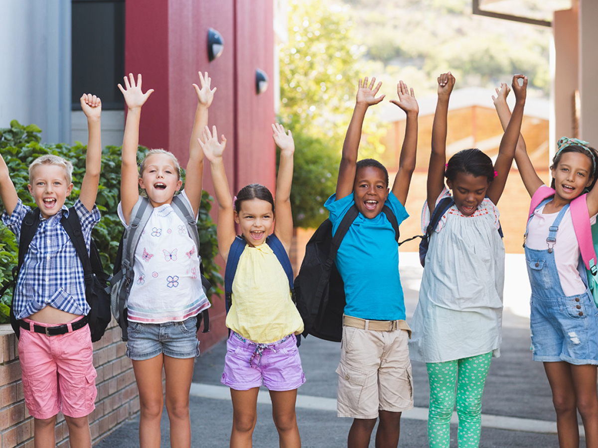 Six children with backpacks raise their hands and smile while standing outdoors in a row.