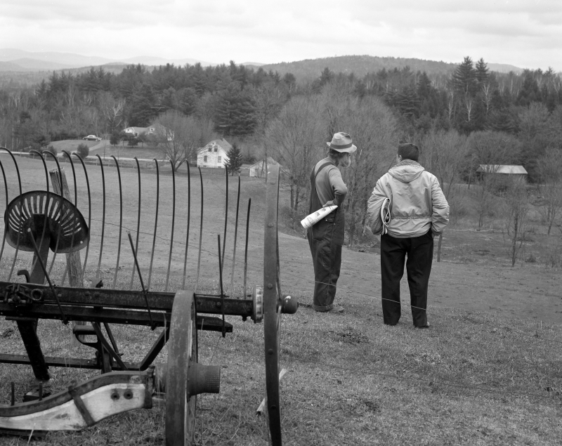 Two men stand on a field overlooking a rural landscape with houses and trees in the background. An old piece of farming equipment is in the foreground.