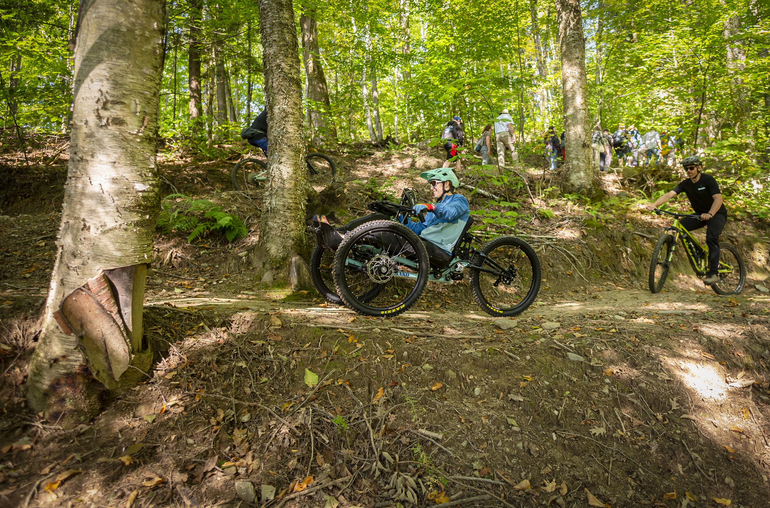 A group of cyclists, including a rider on an adaptive mountain bike, navigate a dirt trail through a dense forest under bright sunlight.