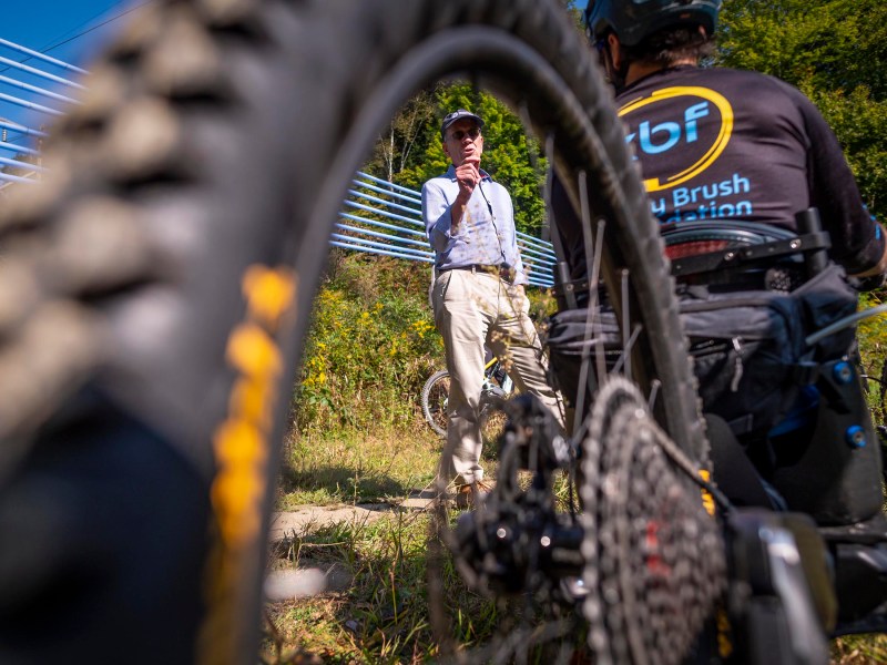 A man stands outdoors speaking to mountain bikers, framed through the bike tires, with greenery and a blue fence in the background.