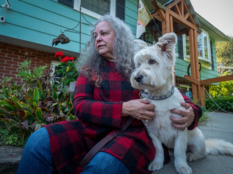 A person with long gray hair sits on the front steps of a house, holding a white dog on their lap.