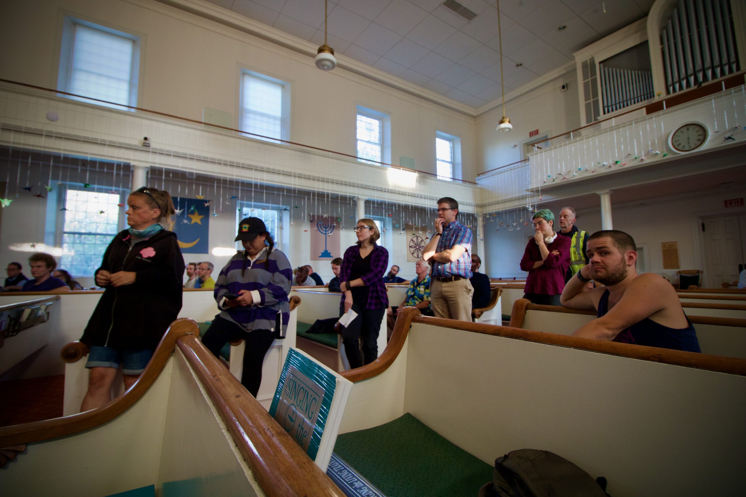 A group of people stands and sits in pews inside a church. The space features a high ceiling, balcony, and various decorations.