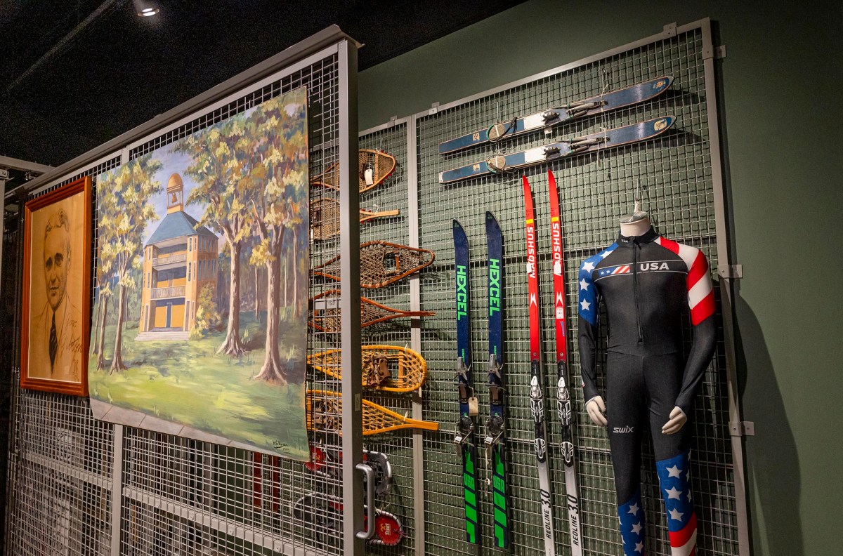A museum display showcases a USA ski suit, several pairs of skis, snowshoes, and a painting of a cabin in the woods, all mounted on a wire grid wall.