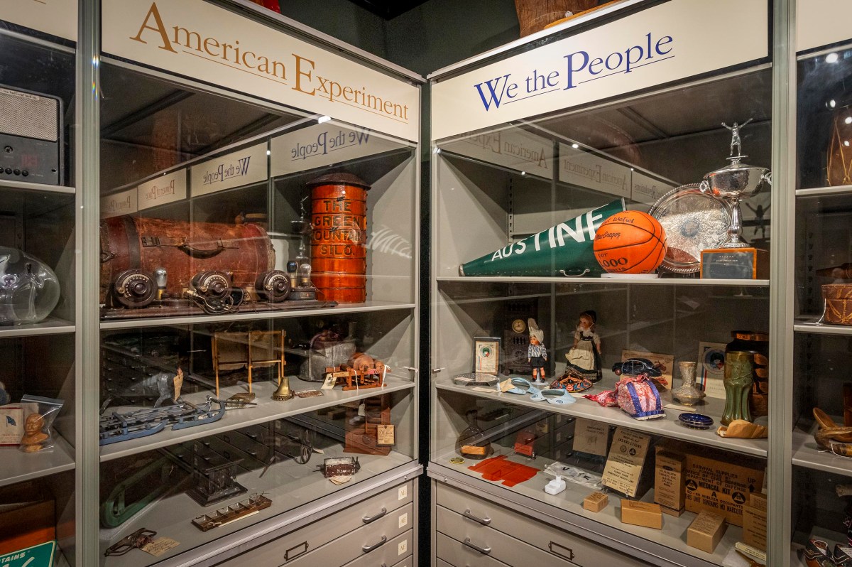 A museum display case features historical artifacts labeled "American Experiment" and "We the People," including a basketball, documents, scale models, and various memorabilia.