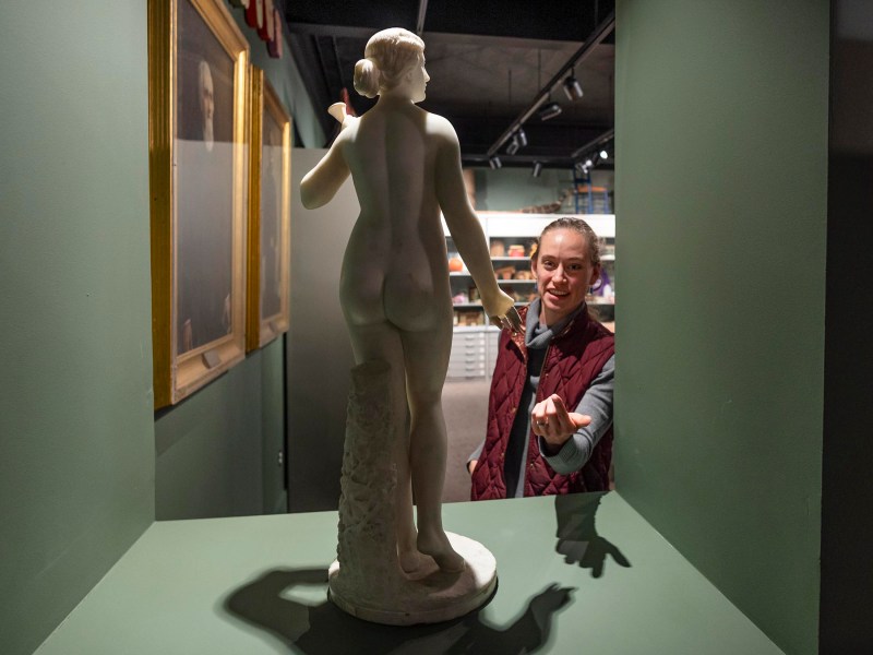 A person gestures towards a classical-style marble statue in an art gallery, with framed artwork on the wall behind them.