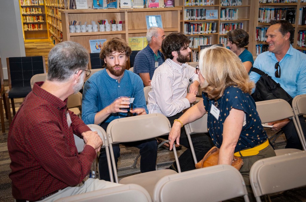 A group of people sit on chairs in a library, engaged in conversation. Shelves of books are seen in the background.