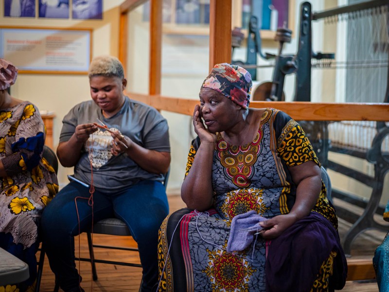 A group of people sitting in a room, with one person knitting and another talking on a cellphone. The setting appears to be casual and relaxed.
