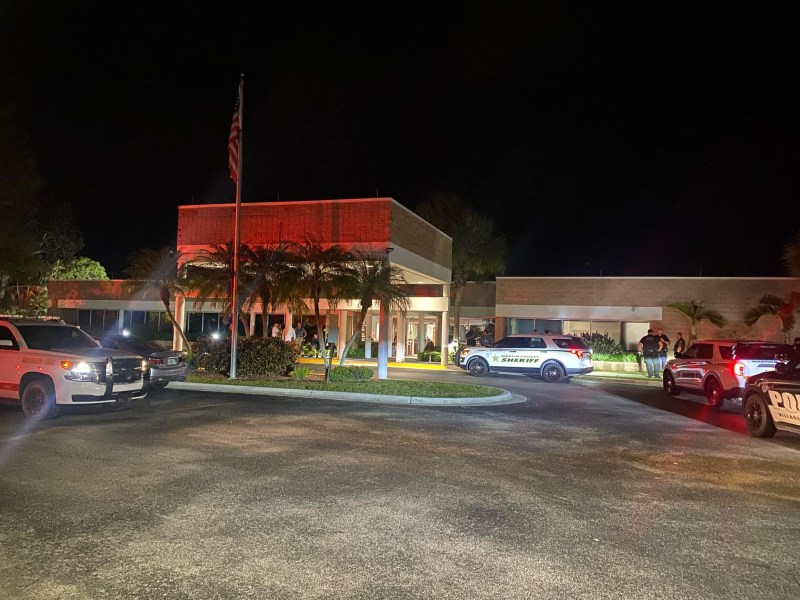 Police vehicles parked outside a building at night. An American flag is visible in front, and the building is illuminated with red lighting. Several people are near the entrance.