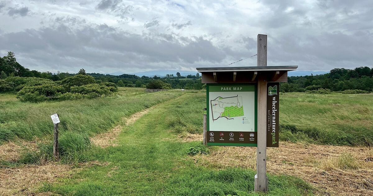 A park sign displaying a map and trail information stands at the entrance to a grassy path, with rolling hills and cloudy skies in the background.