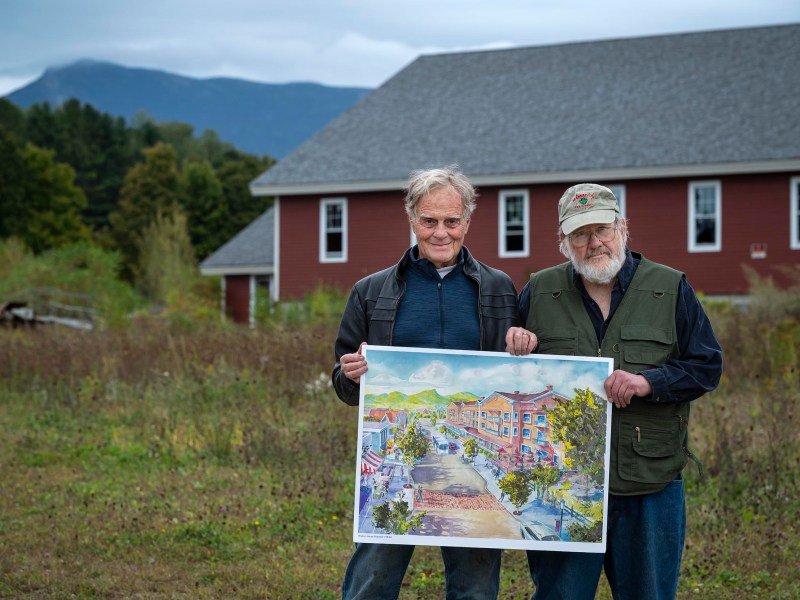 Two men stand outdoors holding a colorful architectural drawing, with a red building and mountainous landscape in the background.