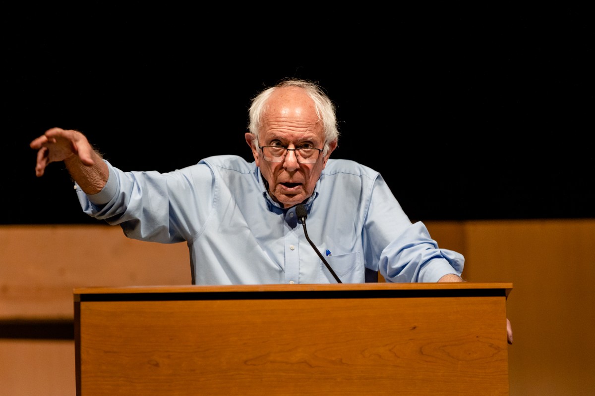 A man with gray hair and glasses speaks passionately at a wooden podium, gesturing with his right hand.