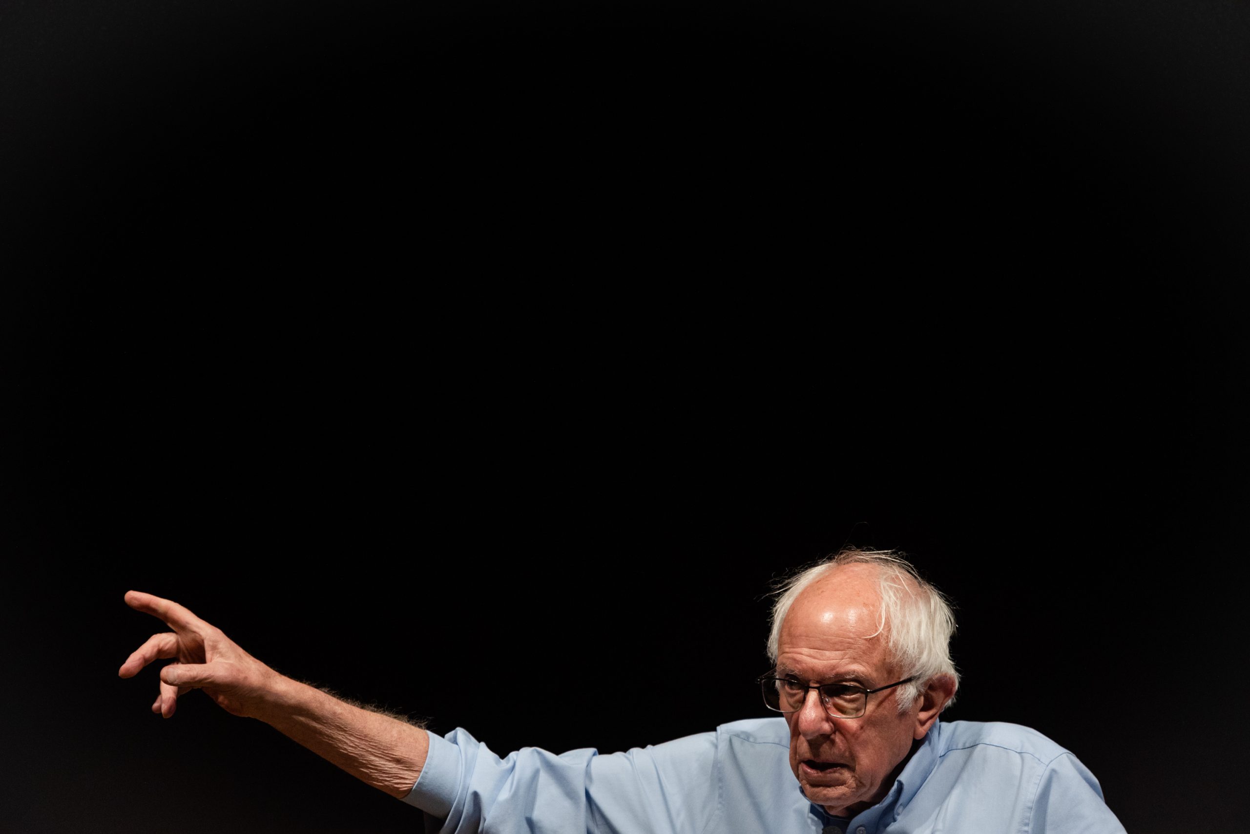 A man gestures while speaking, against a dark background, wearing a light blue shirt and glasses.