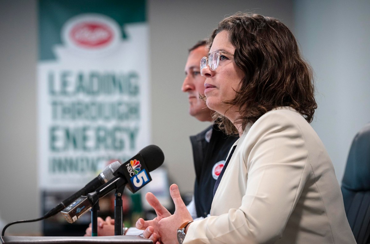 A woman in a white blazer speaks at a podium with microphones, next to a man in a black jacket. A blurred banner in the background reads "Leading Through Energy Innovation.