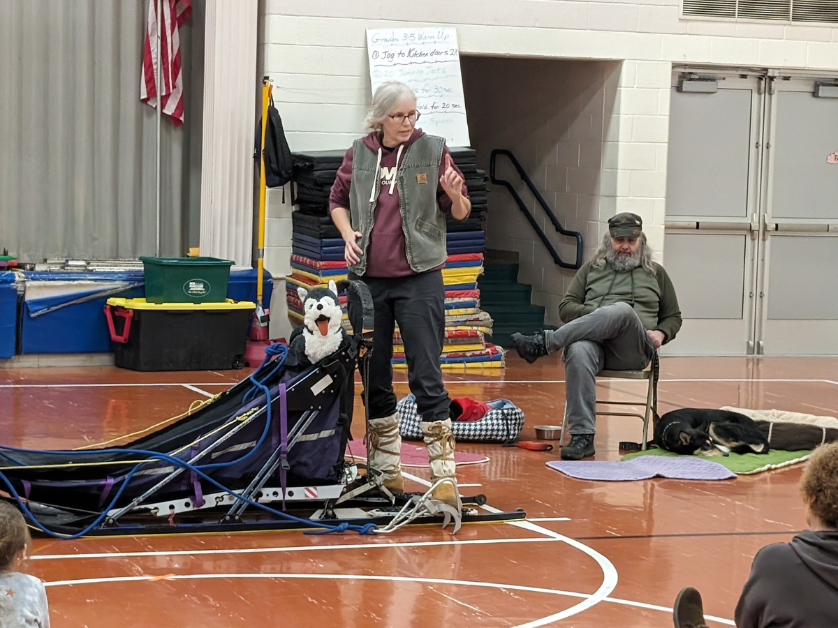 A woman stands next to a sled and speaks, while a man sits nearby with a dog resting beside him in an indoor gym setting.