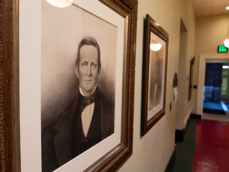 A framed portrait of a man hangs on the wall in a hallway with another framed picture and an open door visible in the background.