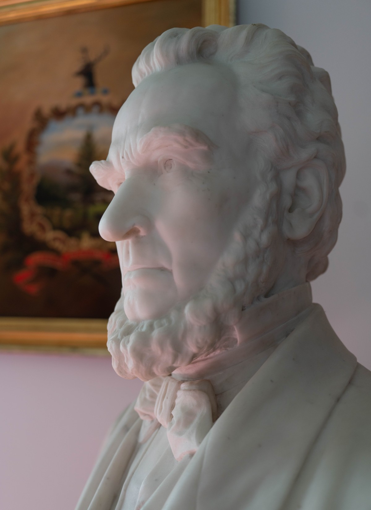 Marble bust of a bearded man in formal attire, set against a blurred background with a framed painting.