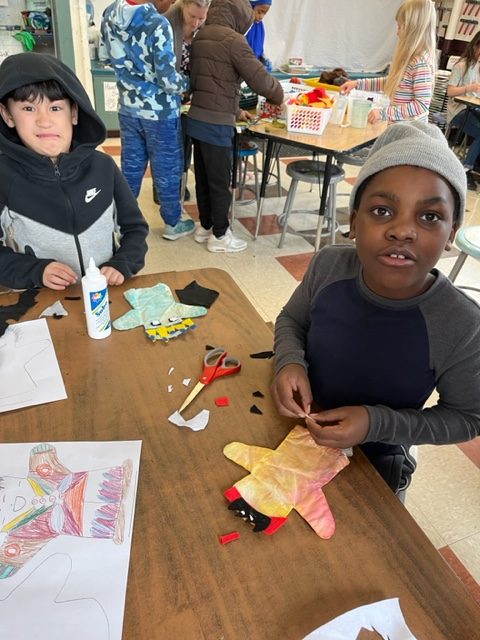 Children engaged in an indoor arts and crafts activity; one child is working with cut fabric, scissors, and glue, while others in the background are working on similar projects.