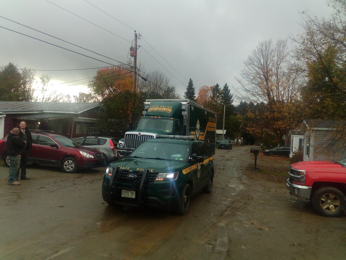 A police SUV and a large green truck are on a muddy road with parked cars and onlookers nearby under a cloudy sky.