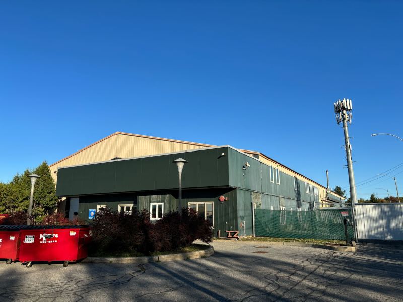 Industrial building with a green exterior and a tan roof. Red dumpsters are in front, and a cell tower stands nearby. Clear blue sky above.
