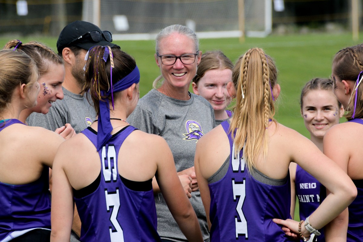 A coach stands smiling among a group of female lacrosse players wearing purple jerseys with numbers, engaged in a casual team huddle on a field.