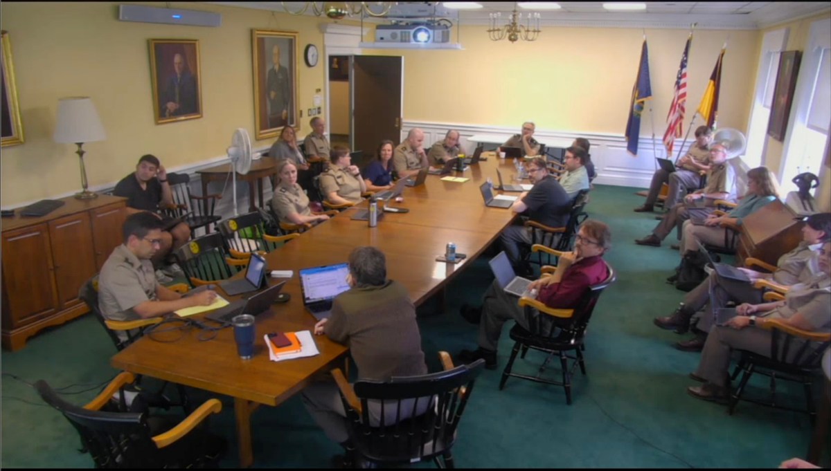 People in uniformed attire sit around a long conference table in a meeting room with portraits on the walls and flags in the corner.