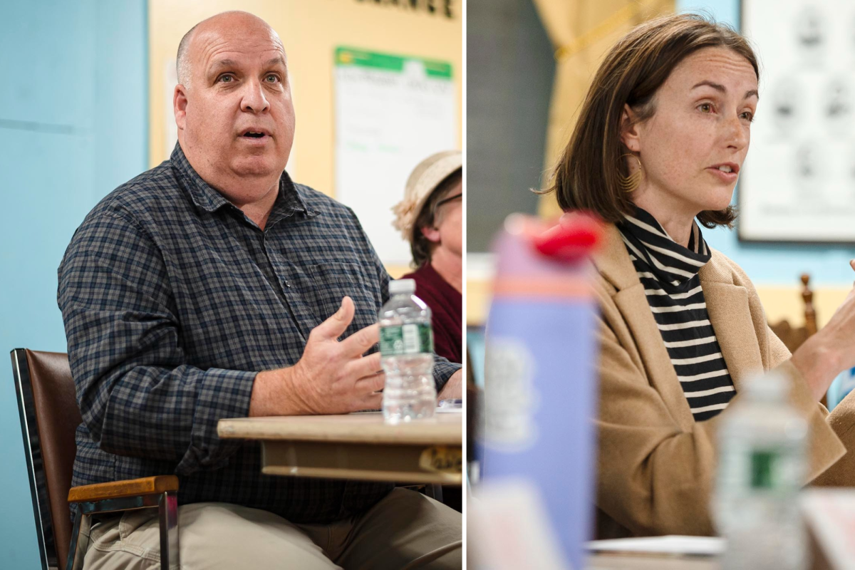 Two people seated at a table, engaged in discussion. One man on the left wears a checkered shirt, and a woman on the right wears a striped top and tan coat. Water bottles are on the table.
