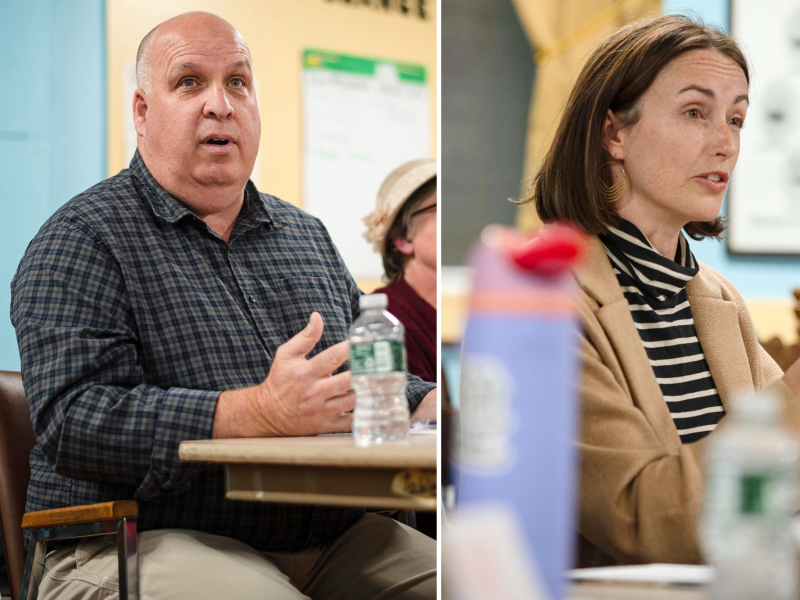 Two people seated at a table, engaged in discussion. One man on the left wears a checkered shirt, and a woman on the right wears a striped top and tan coat. Water bottles are on the table.