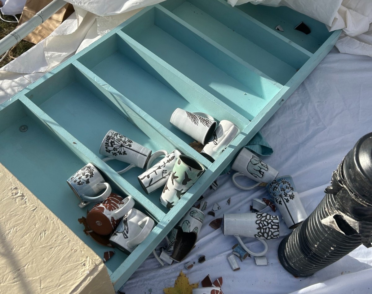 Broken ceramic mugs on the ground beside an overturned light blue shelf. Some mugs are intact, while others are shattered.