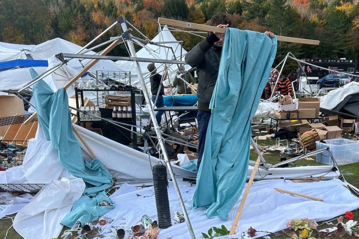 A person is disassembling a collapsed tent frame surrounded by scattered fabric, boxes, and items on the ground at an outdoor setting.