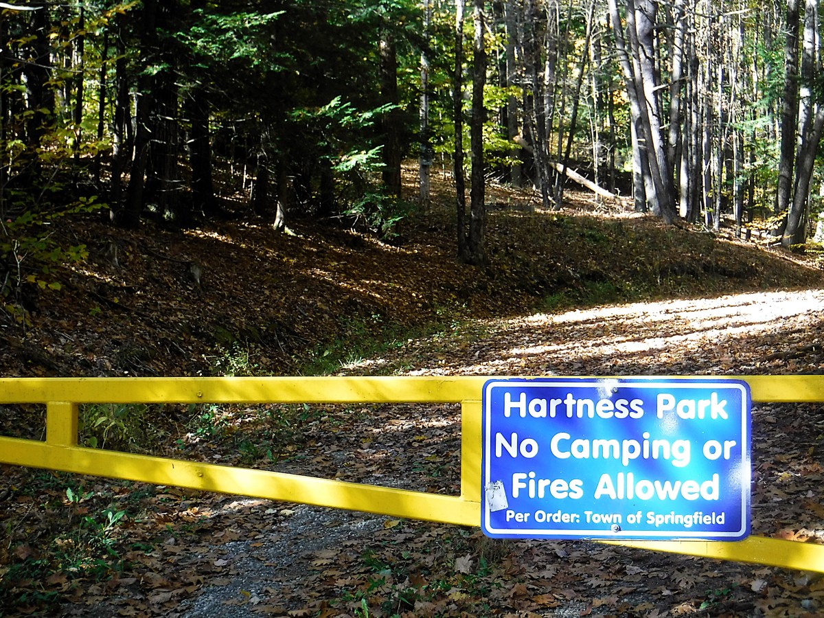 A trail entrance in a forest with a yellow gate. A sign reads, "Hartness Park. No camping or fires allowed. Per Order, Town of Springfield.
