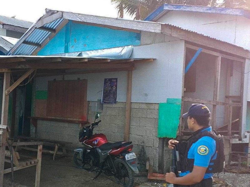 A police officer holding a weapon stands in front of a building with a metal roof and two parked motorcycles.
