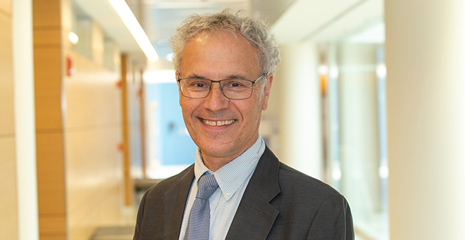 A smiling man with gray hair and glasses, wearing a suit and tie, stands in a brightly lit hallway.