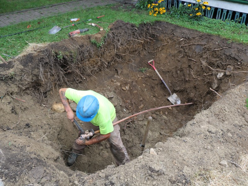 A worker in a blue helmet and yellow shirt digs a deep hole in the ground with a shovel, surrounded by dirt and roots. Garden tools are scattered nearby.