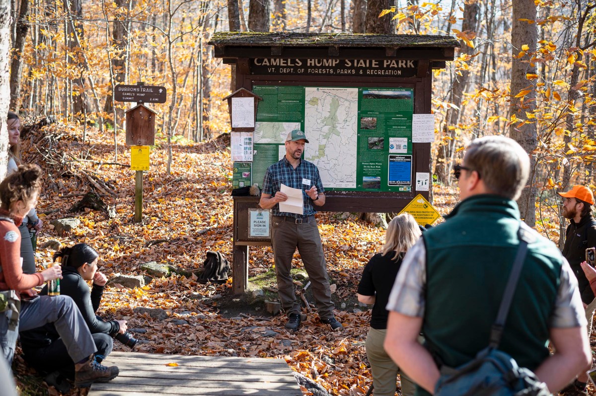 A person speaks to a group in front of a large information board at Camels Hump State Park, surrounded by autumn foliage.