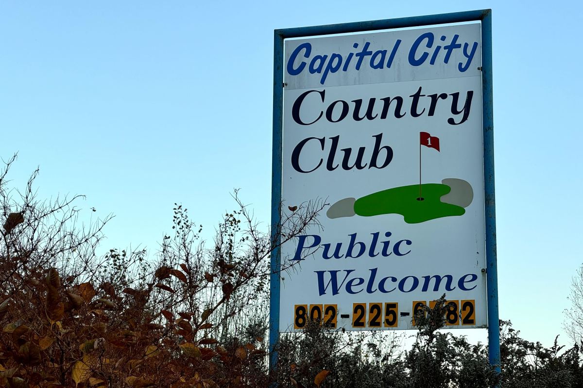 Sign for Capital City Country Club with golf green graphic. Text reads: "Public Welcome" and includes a phone number. Surrounded by plants and a clear sky in the background.