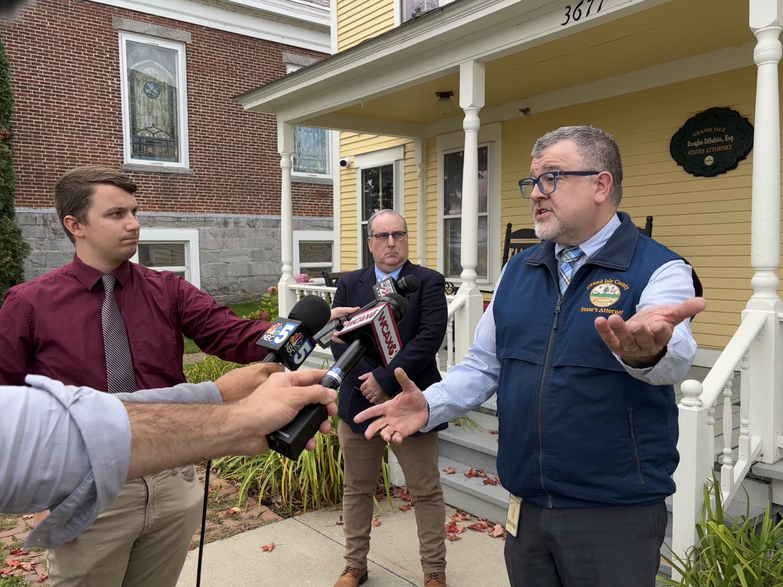 A man in a vest speaks to reporters holding microphones outside a building with a porch. Two other men stand nearby, one holding a notebook.