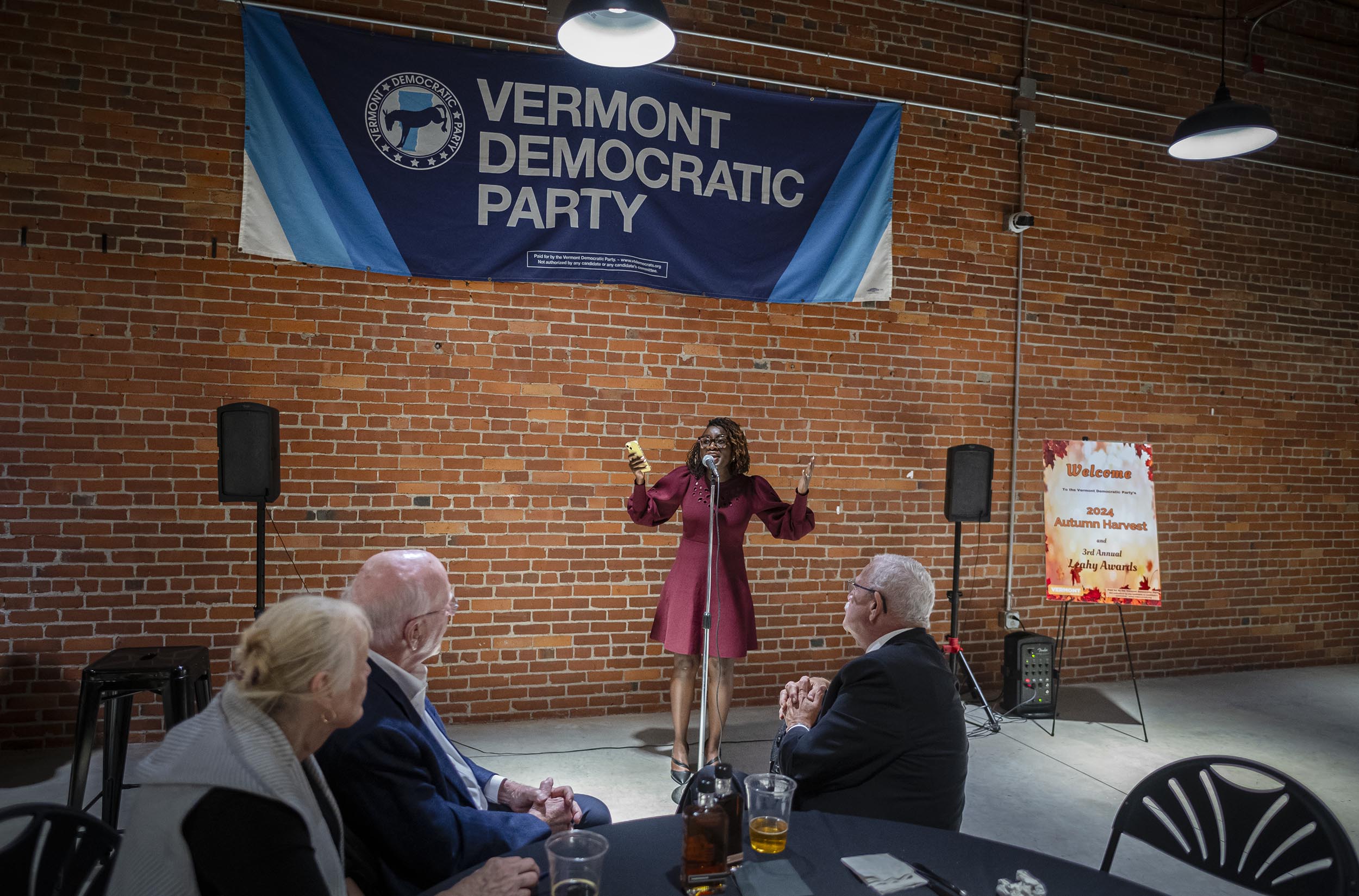 A person speaks on stage at a Vermont Democratic Party event, with an audience seated at tables and a brick wall in the background.