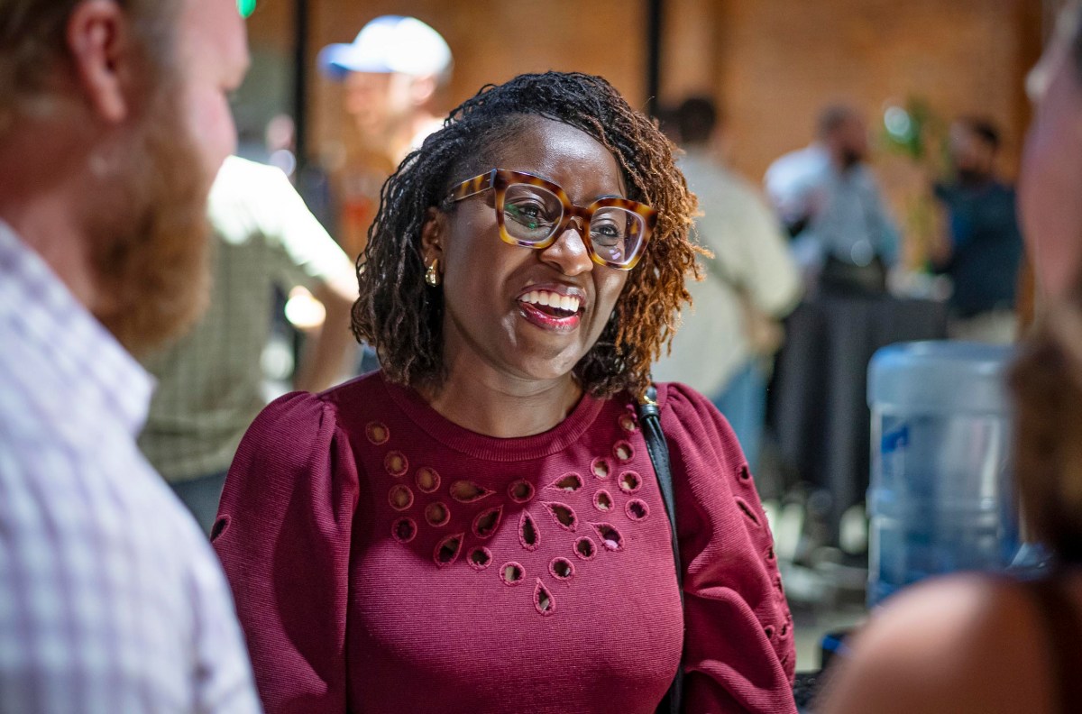 A woman wearing glasses and a maroon top smiles while talking to two people at an indoor event.