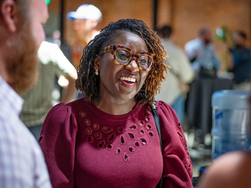 A woman wearing glasses and a maroon top smiles while talking to two people at an indoor event.