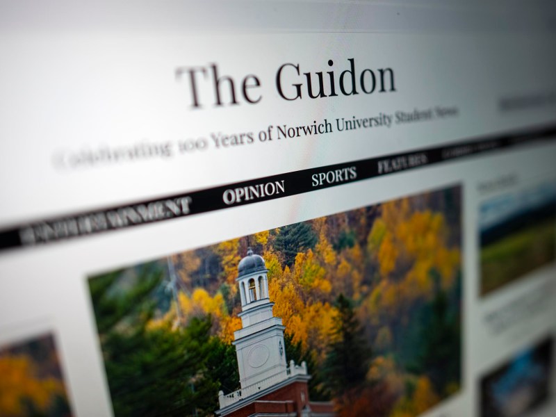 A website screenshot of "The Guidon," celebrating 100 years of Norwich University student news, featuring a photo of a building surrounded by autumn trees.