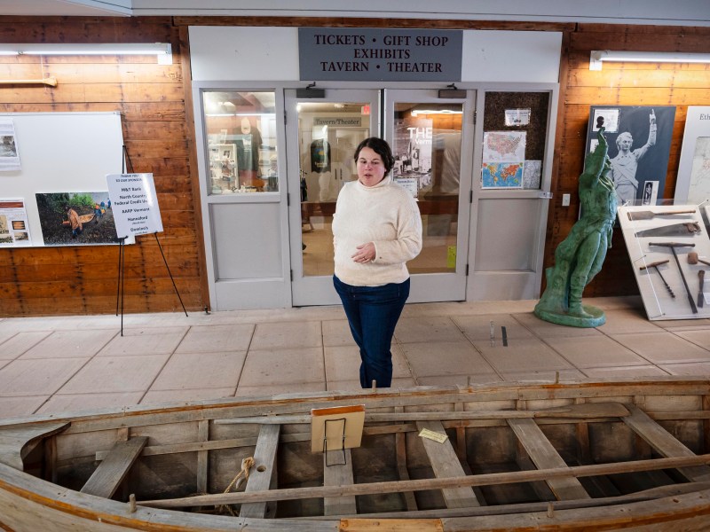 A person stands in a museum near a wooden boat exhibit. Signs for tickets, gift shop, tavern, and theater are visible in the background.
