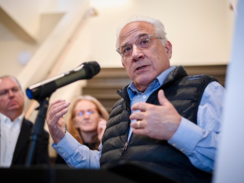 An older man in glasses and a vest speaks at a microphone, gesturing with his hand. Two people in the background listen attentively.