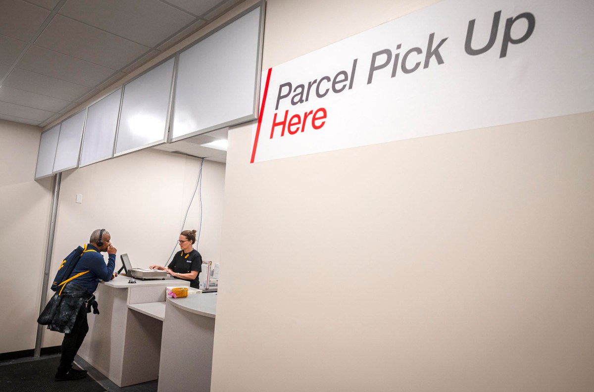 A person interacts with an employee at a parcel pick-up counter. A sign reads "Parcel Pick Up Here.