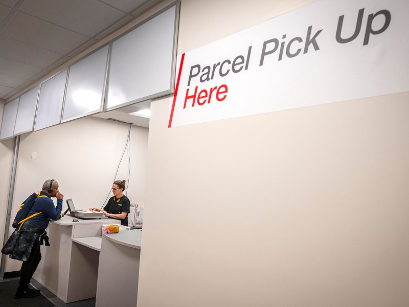 A person interacts with an employee at a parcel pick-up counter. A sign reads "Parcel Pick Up Here.