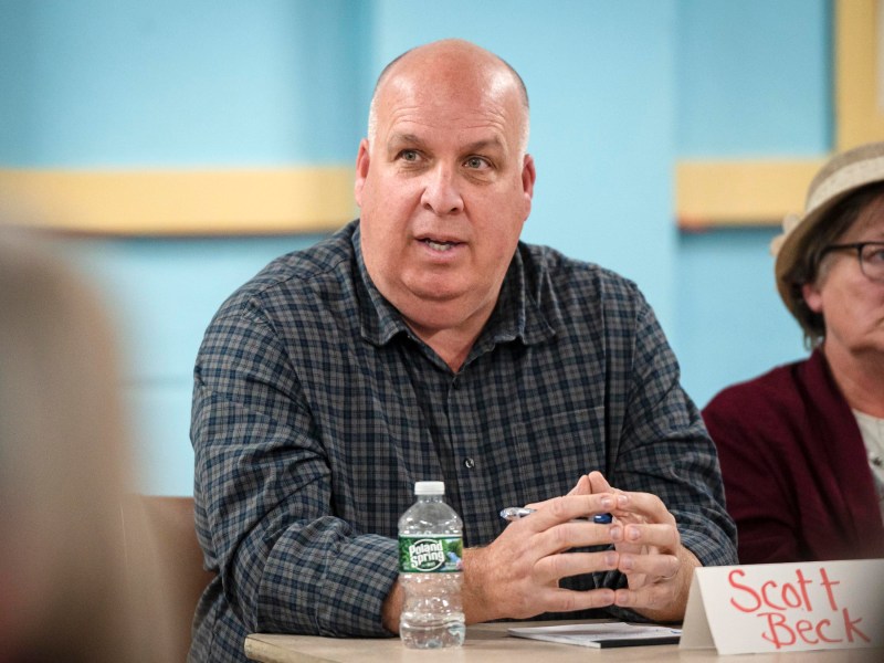 A man sits at a table with a name card reading "Scott Beck," speaking, and gesturing with hands. A water bottle and another person are visible nearby.