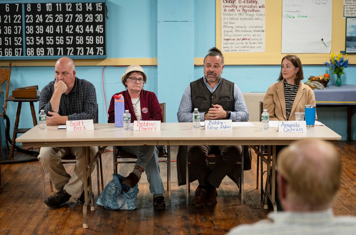 Four people are seated at a table with name cards in a community hall, participating in a panel discussion. Audience members are visible in the foreground.
